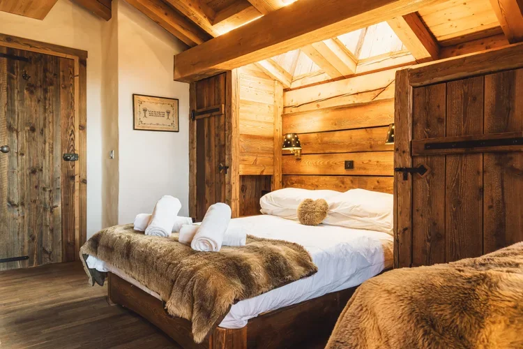 Cozy rustic bedroom with wooden walls, a bed with white linens and fur throws, pillows, and rolled towels, under a skylight in a cabin style interior.