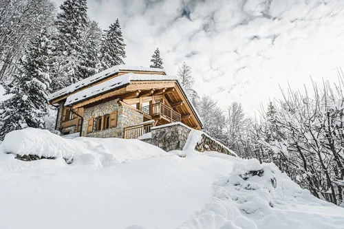 A snow-covered wooden cabin on a hillside surrounded by snow and trees, with a cloudy sky overhead.