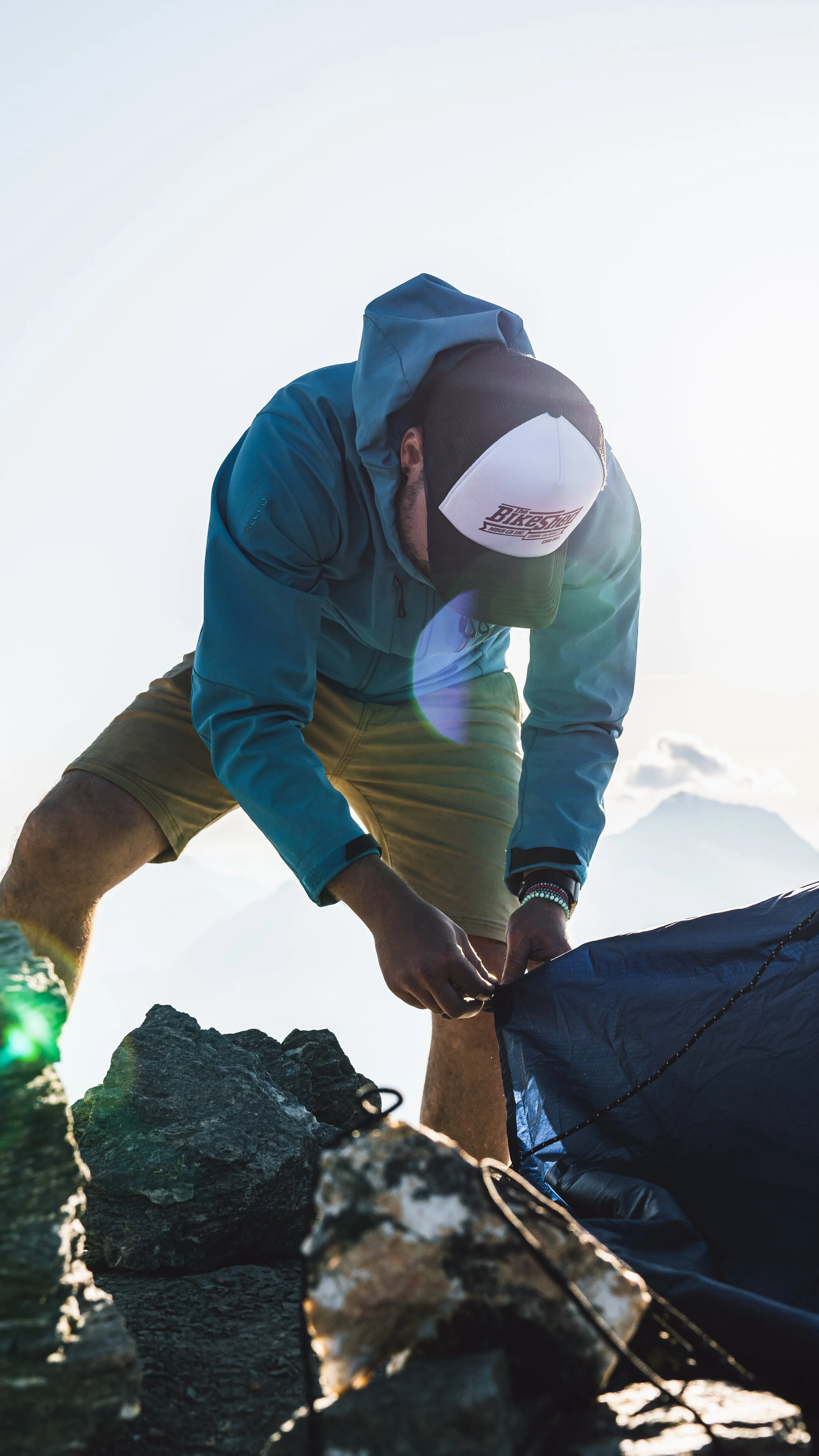 A man adjusting camping gear on rocky terrain outdoors with mountain and sky background