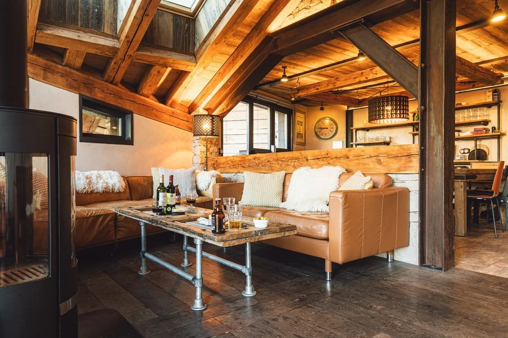 Cozy living room with wooden beams, leather sofas with furry pillows, a rustic coffee table with drinks, and a bookshelf, featuring warm lighting and a rustic aesthetic.