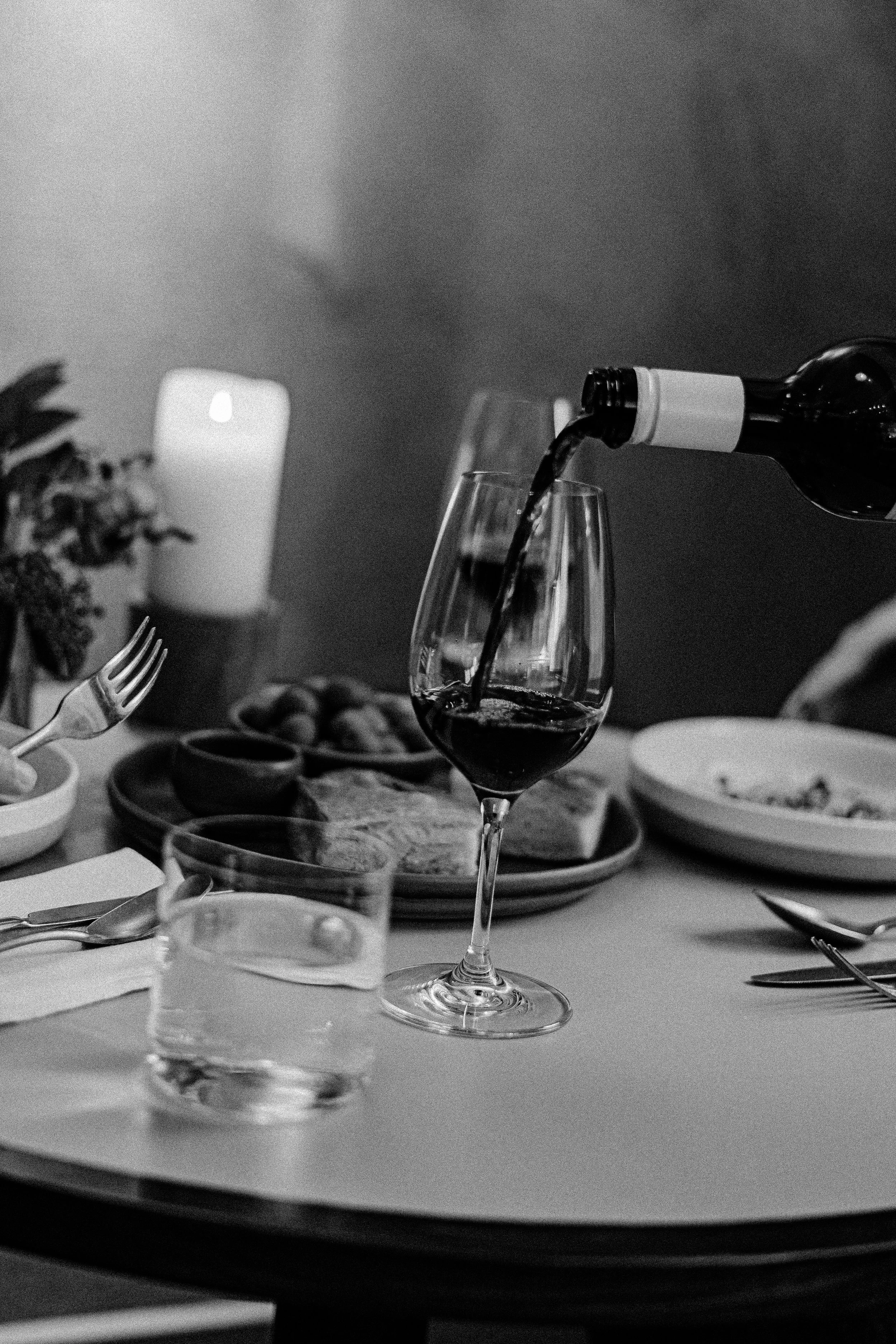 A black-and-white photo of wine being poured into a glass at a dinner table with plates of food, cutlery, a glass of water, a candle, and a plant in the background.