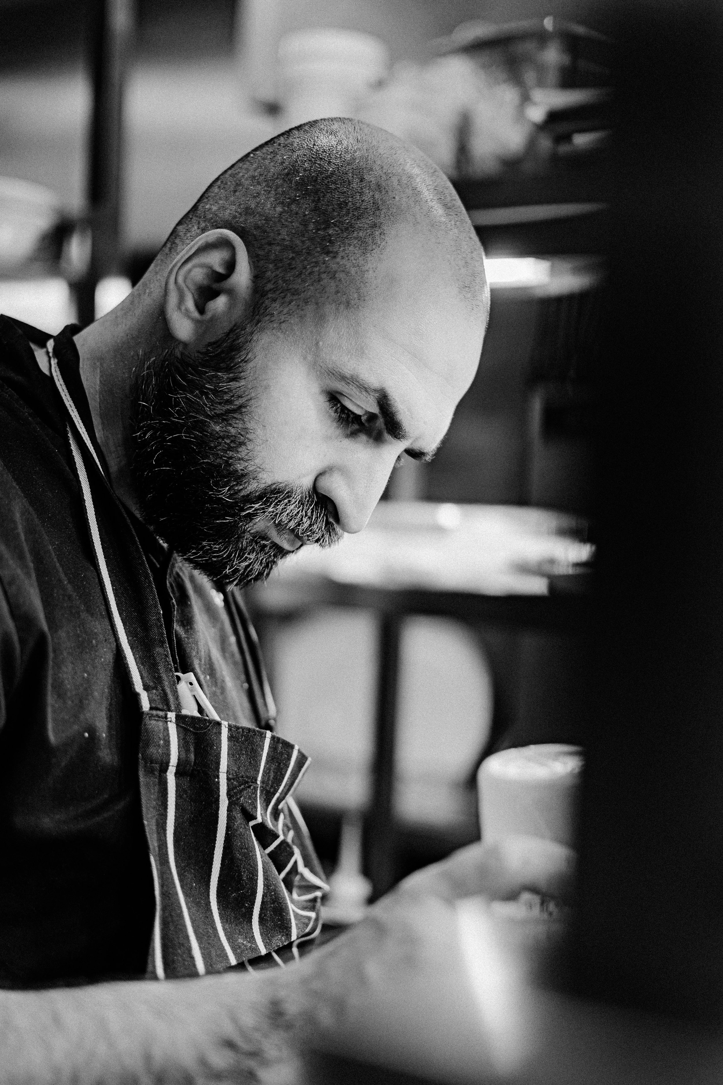 Black and white photo of a man with a beard and shaved head, focused on working in a kitchen, wearing an apron.