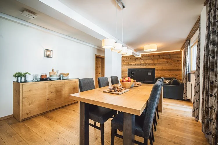 Dining room with a wooden table, six black chairs, a sideboard with plants and dishes, and a wood-paneled wall with a TV, featuring modern lighting and curtains.
