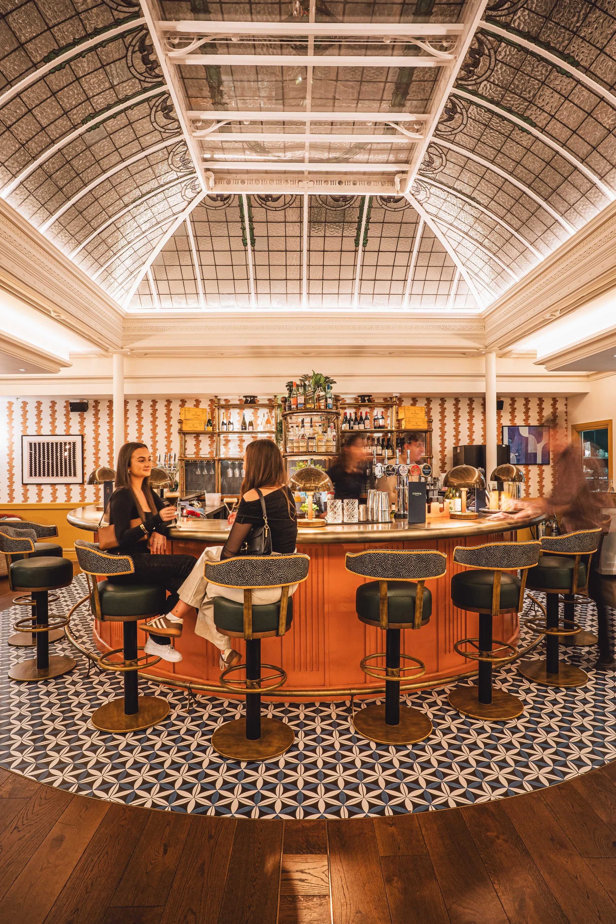 A bar with a rounded counter, two women sitting and talking, and a decorative tiled floor, inside a building with a glass ceiling.