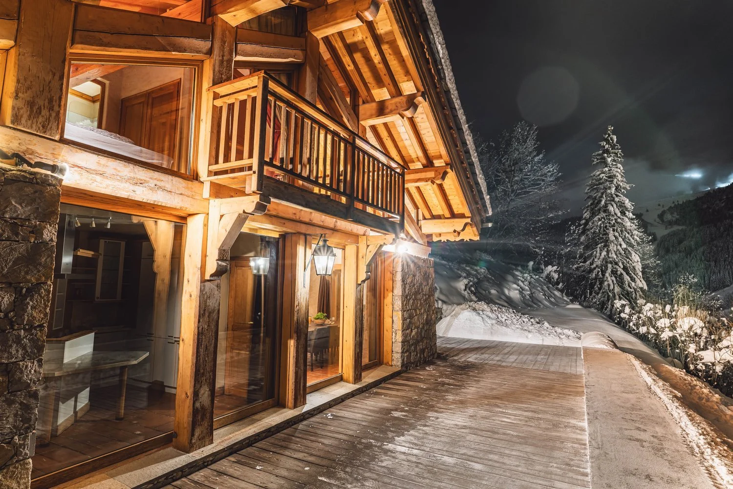 Exterior view of a wooden chalet at night, illuminated by exterior lights, with snow on the ground and a snow-covered pine tree in the background.