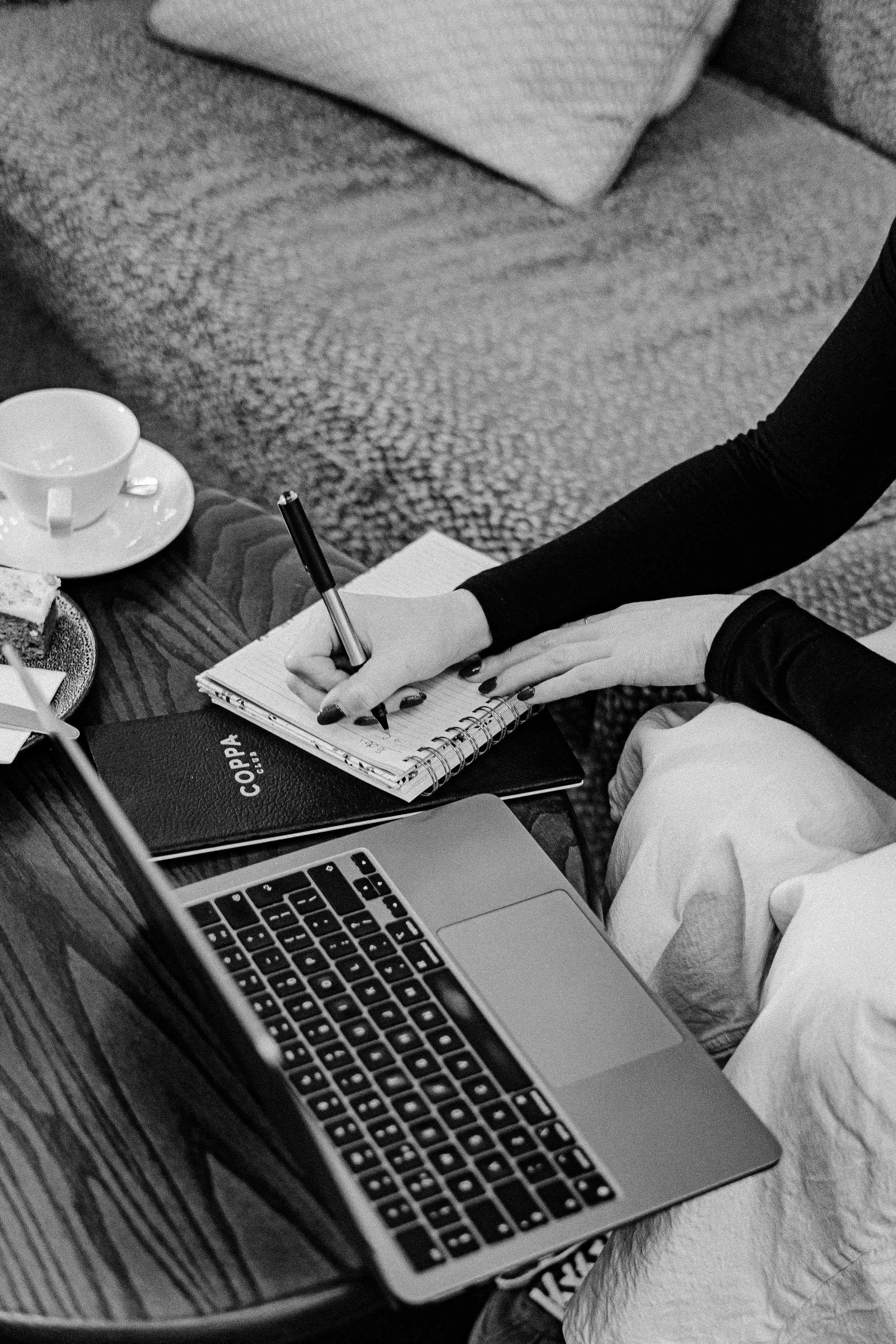 A person with painted nails writing in a small spiral notebook on a coffee table beside an open laptop, a cup of coffee, and some snacks.