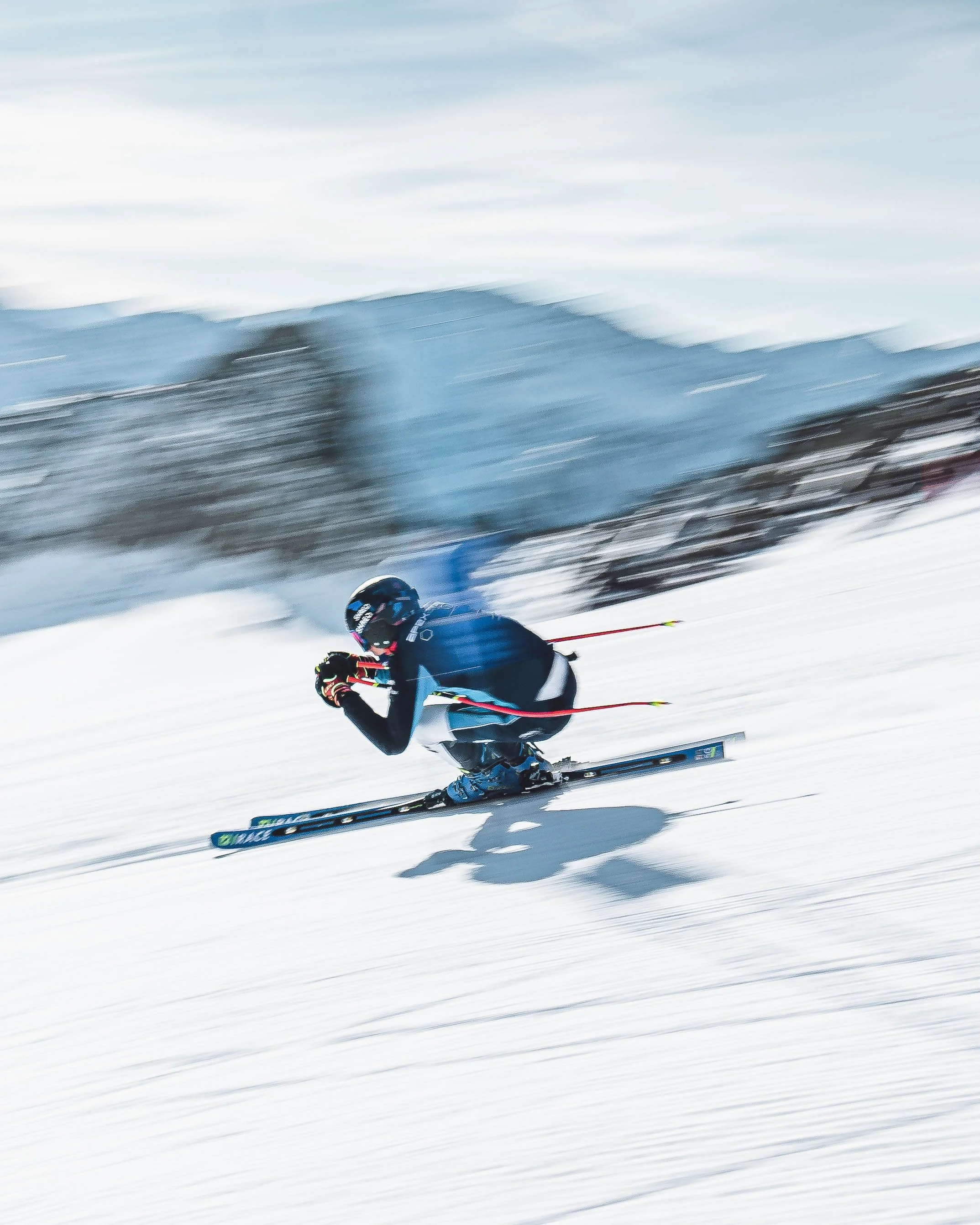 A skier in a black helmet and blue jacket racing down a snowy slope with a blurred background of mountains and ski tracks.