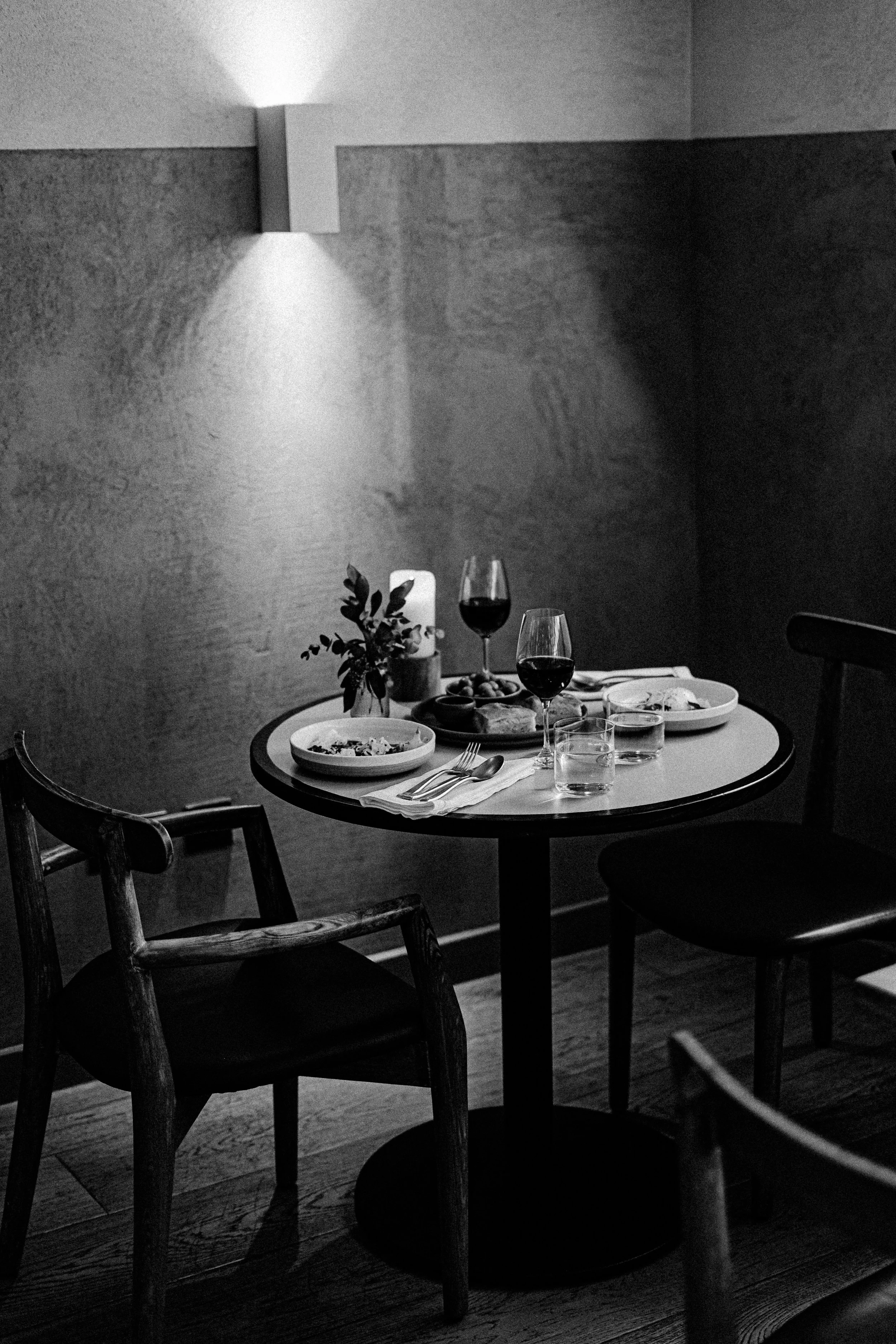 A black and white photo of a round dining table with two glasses of red wine, plates of food, a lit candle, and a small plant, surrounded by chairs, illuminated softly on a textured wall.