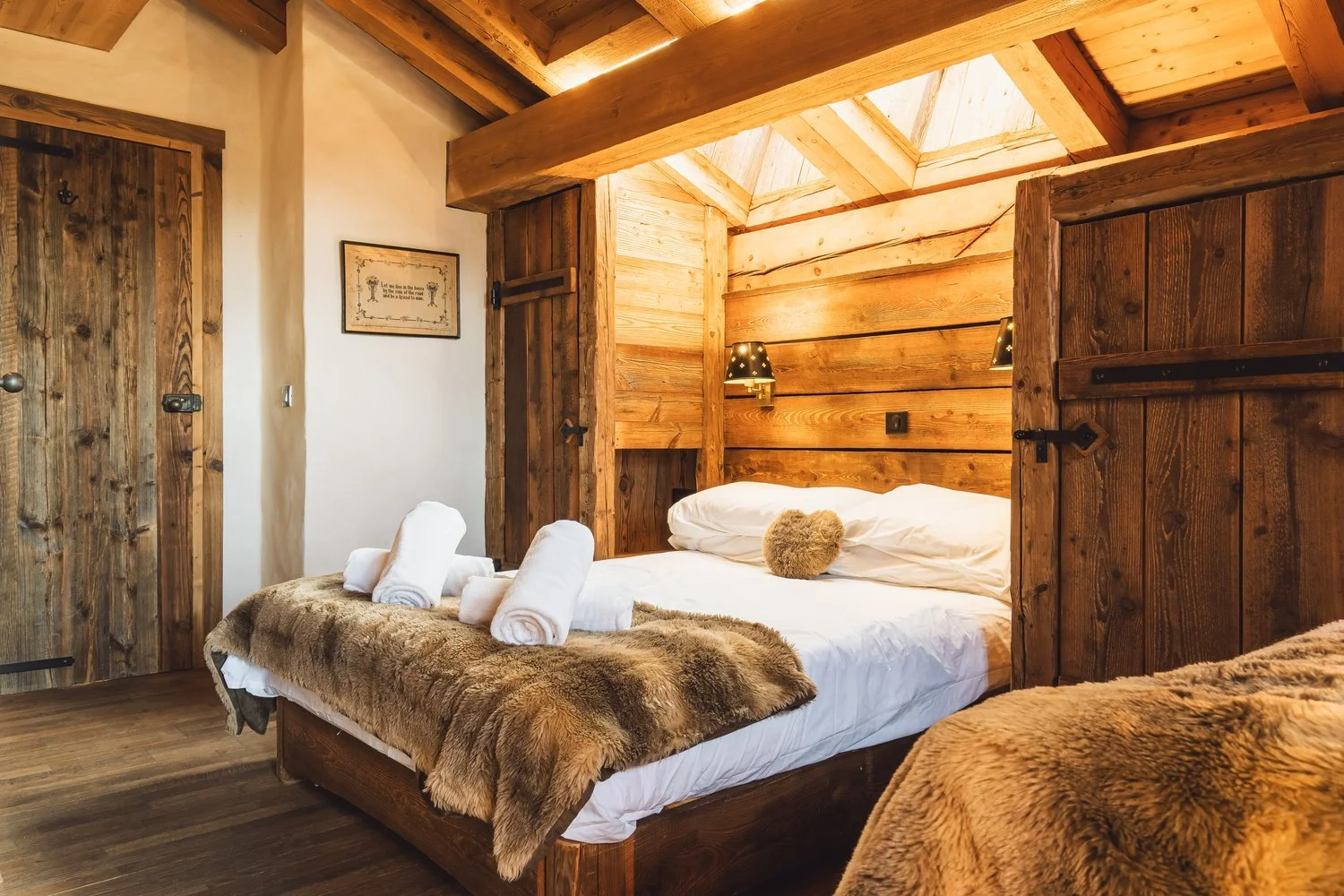 A rustic bedroom with a wooden bed frame and walls, featuring a white bed with rolled towels and a fur blanket, two black wall-mounted lamps, and a skylight ceiling