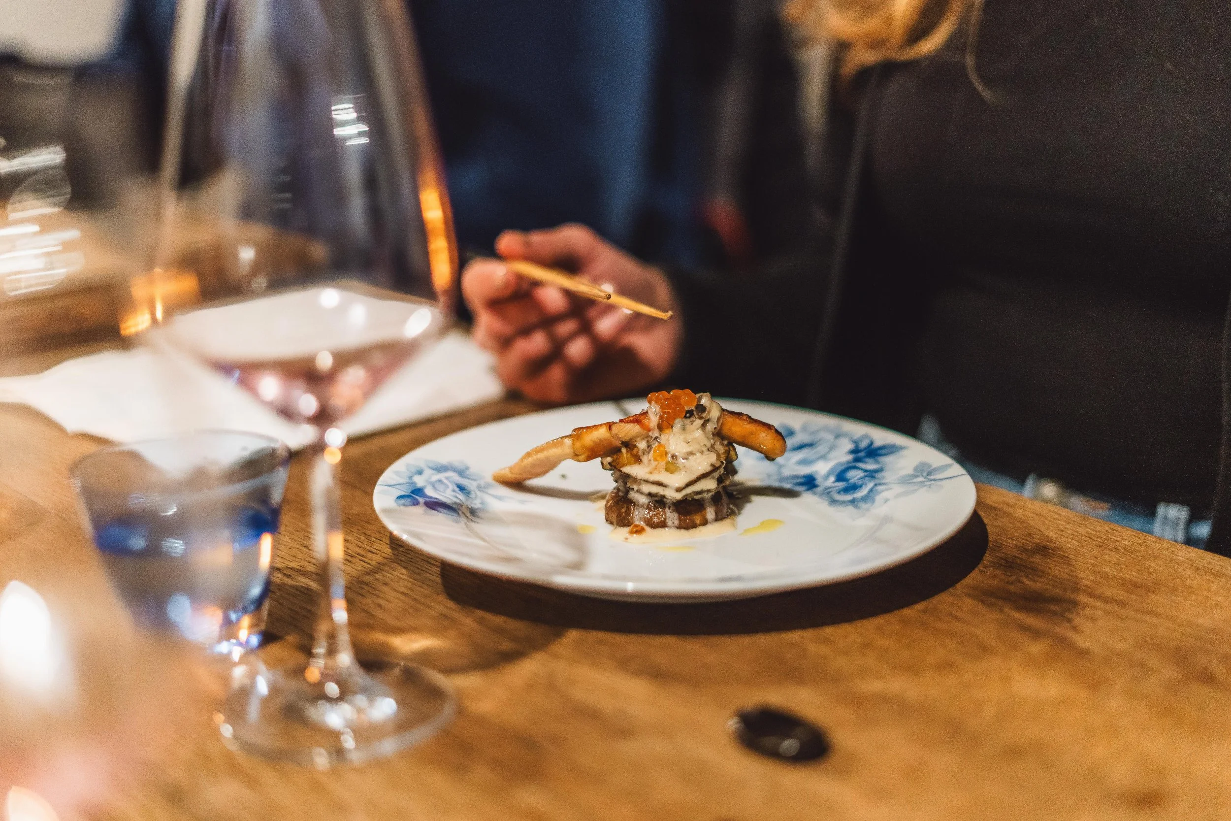 A plated gourmet dish with seafood and caviar, garnished with sauce, in front of a person holding chopsticks, with a glass of white wine and a glass of water on the wooden table.