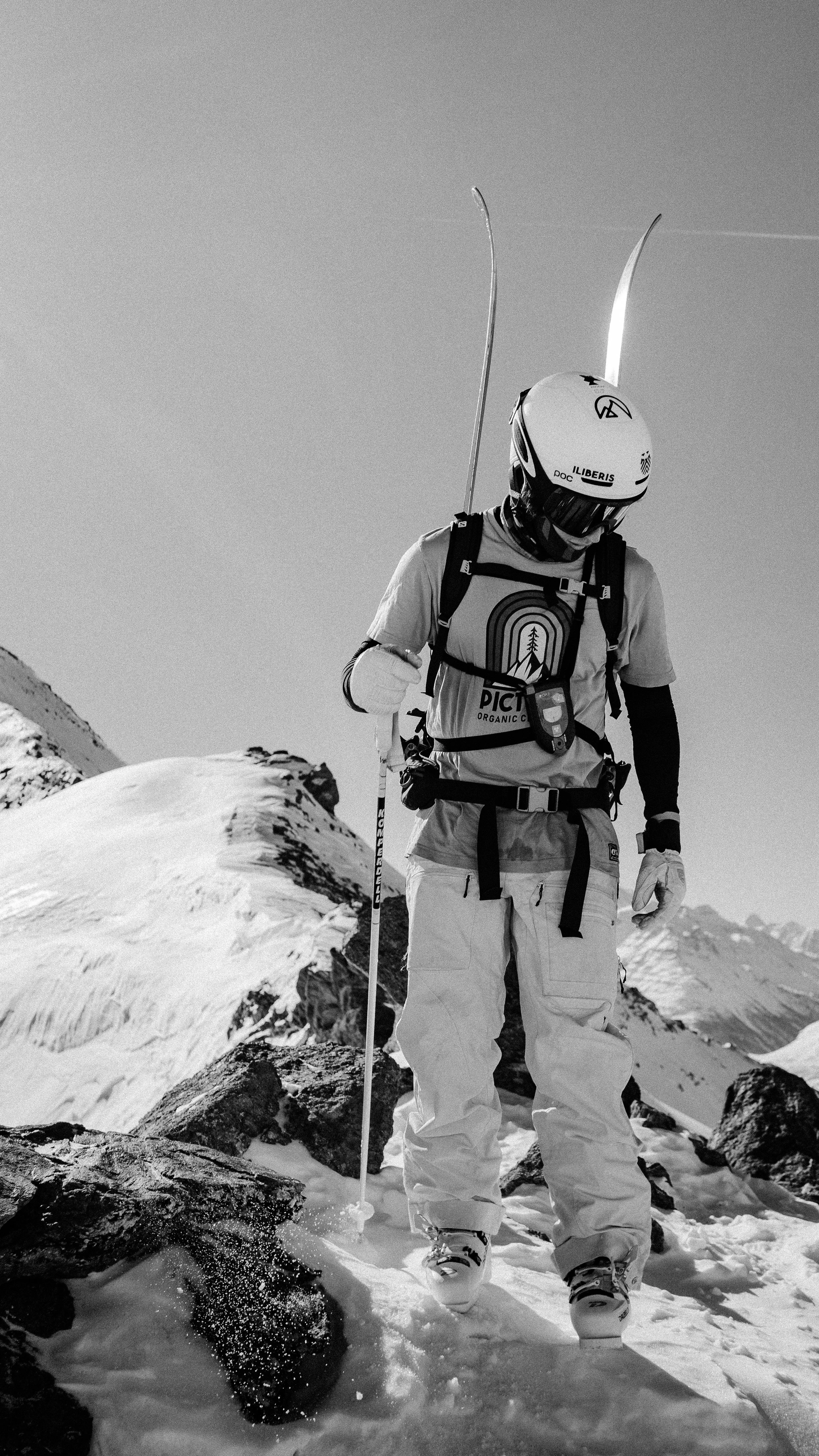 A mountain climber wearing safety gear and snow boots standing on snow-covered terrain with rocky outcroppings and mountain peaks in the background, holding a ski pole with skis on the person's back.
