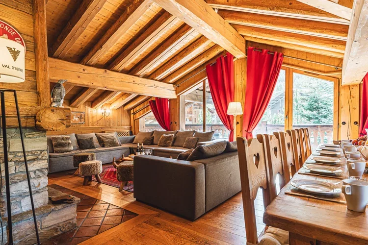 Interior of a cozy wooden cabin dining area with a large sectional sofa, a rustic stone fireplace, a wooden dining table set with plates, cups, and napkins, and large windows with red curtains.