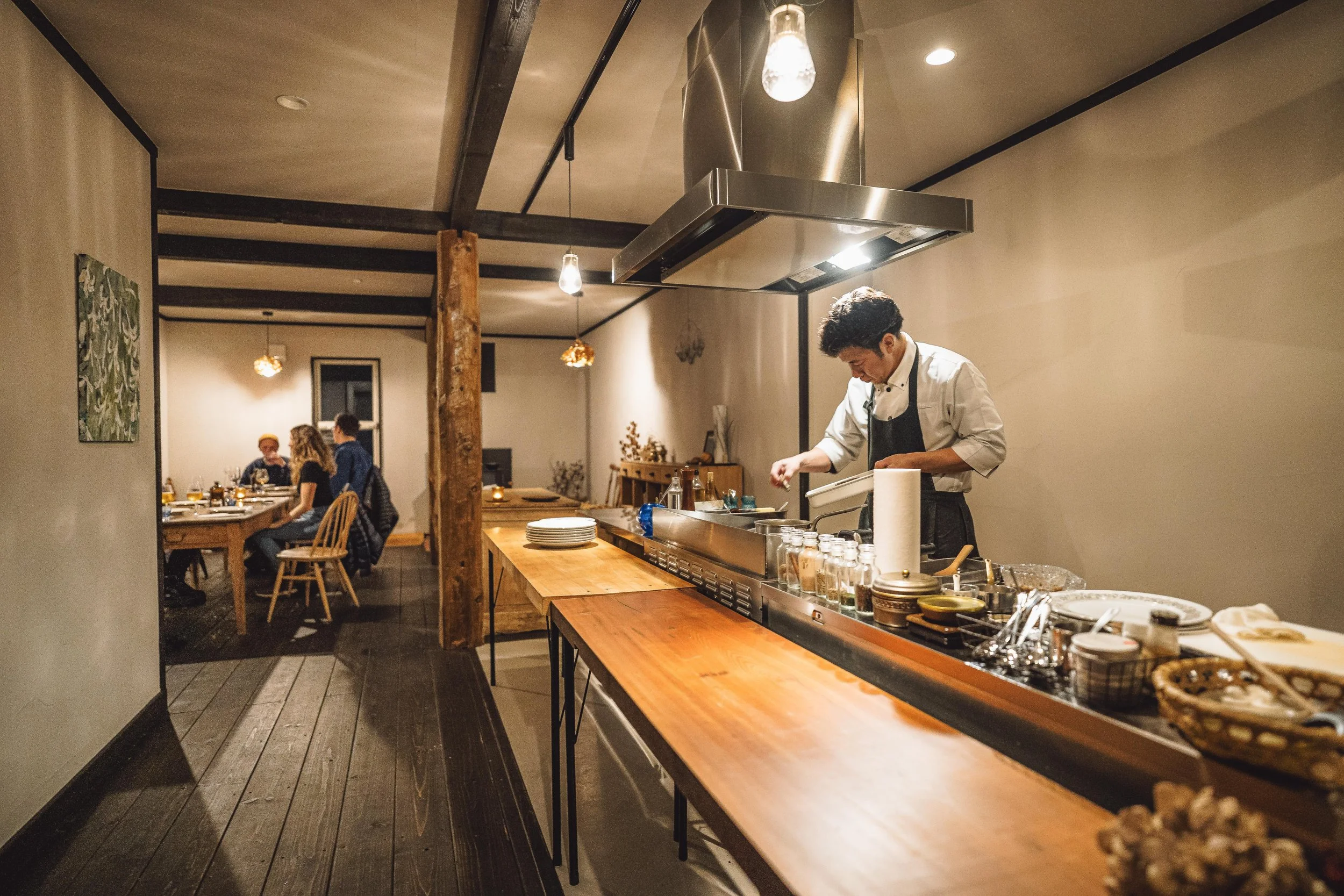 A chef preparing food at a restaurant kitchen counter while customers dine at tables in the background.