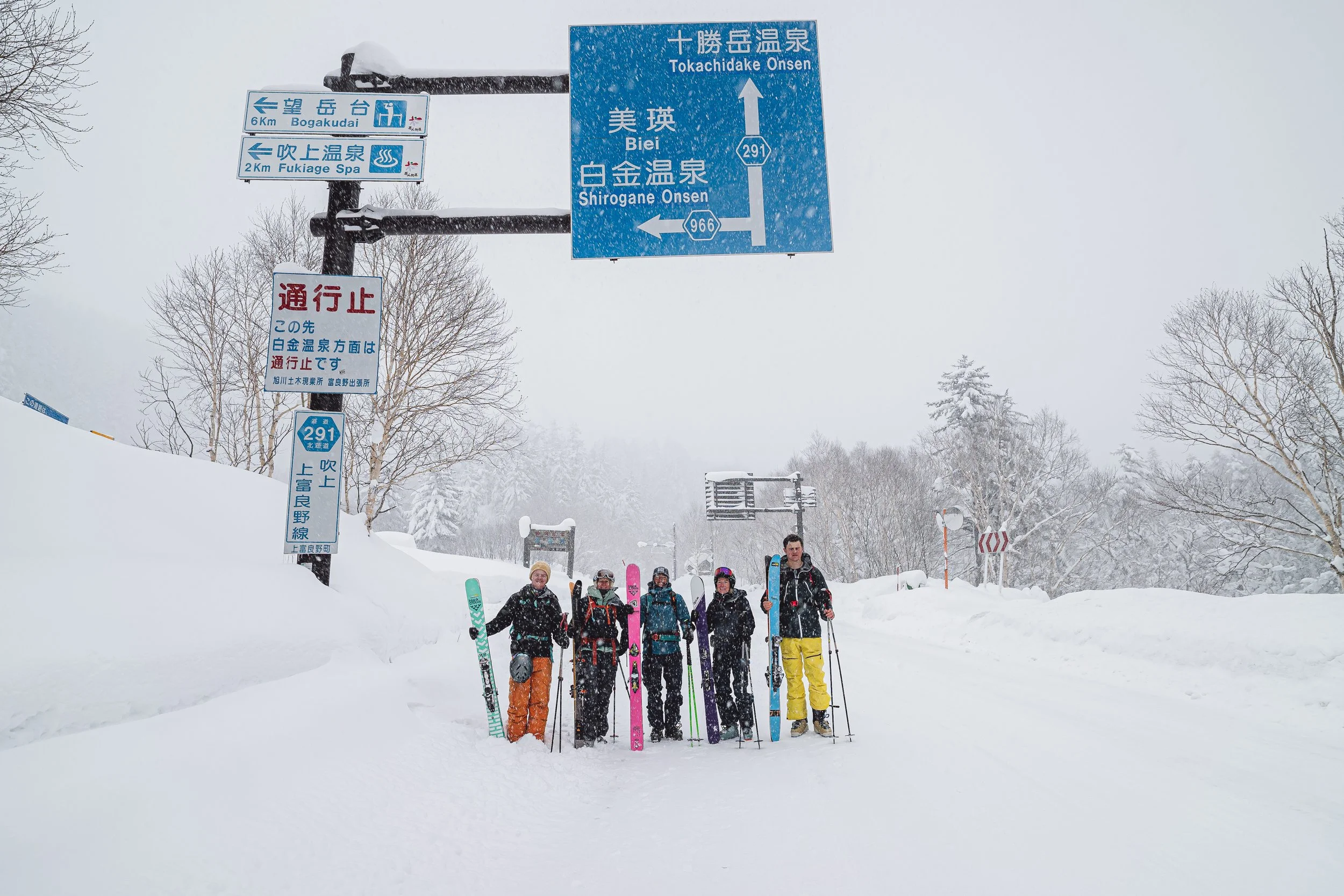 Group of five people standing with ski equipment in a snowy landscape under blue and white directional signs in Japanese, with snow-covered trees and a cloudy sky in the background.