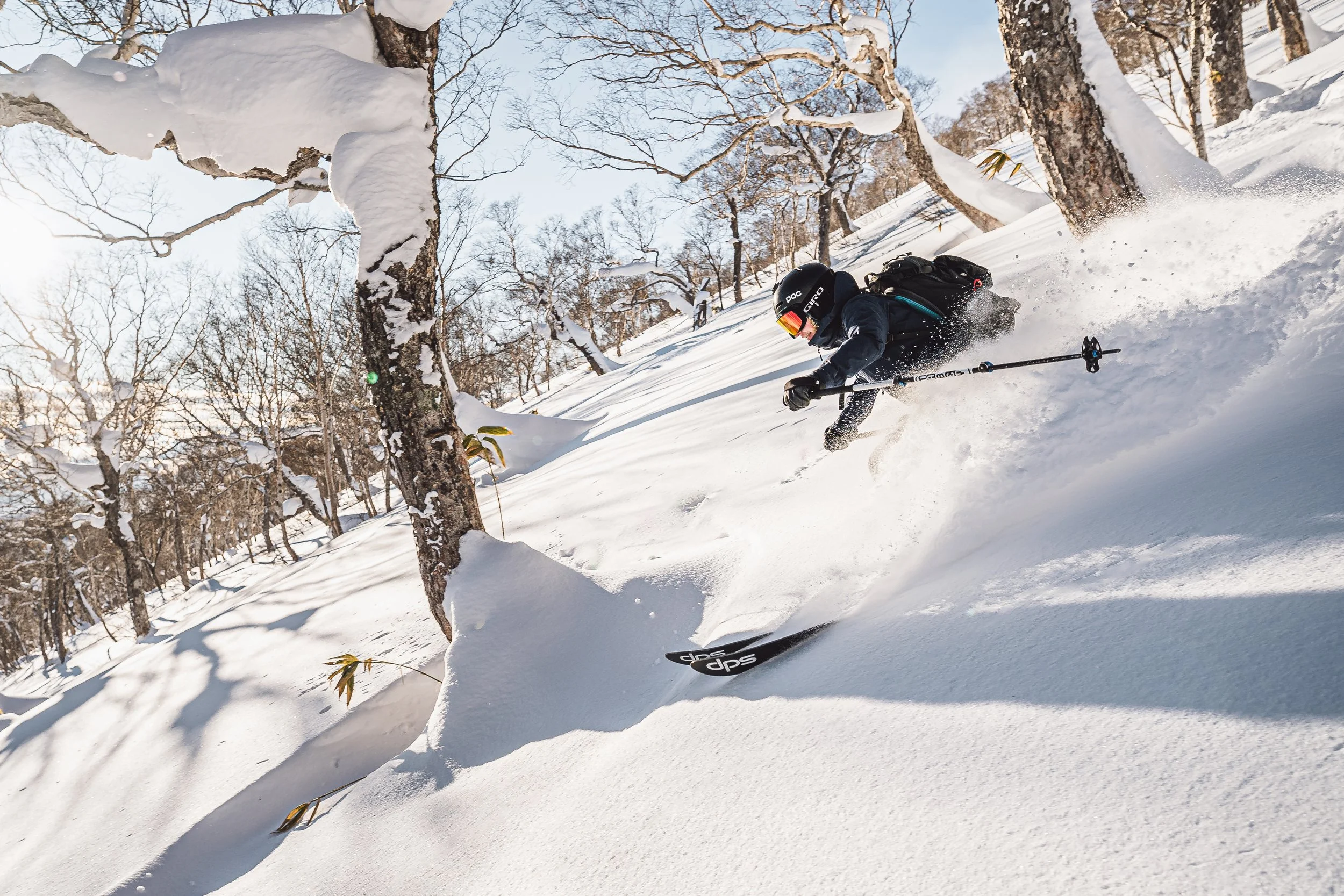 A person skiing down a snow-covered slope with bare trees and a clear sky in the background.