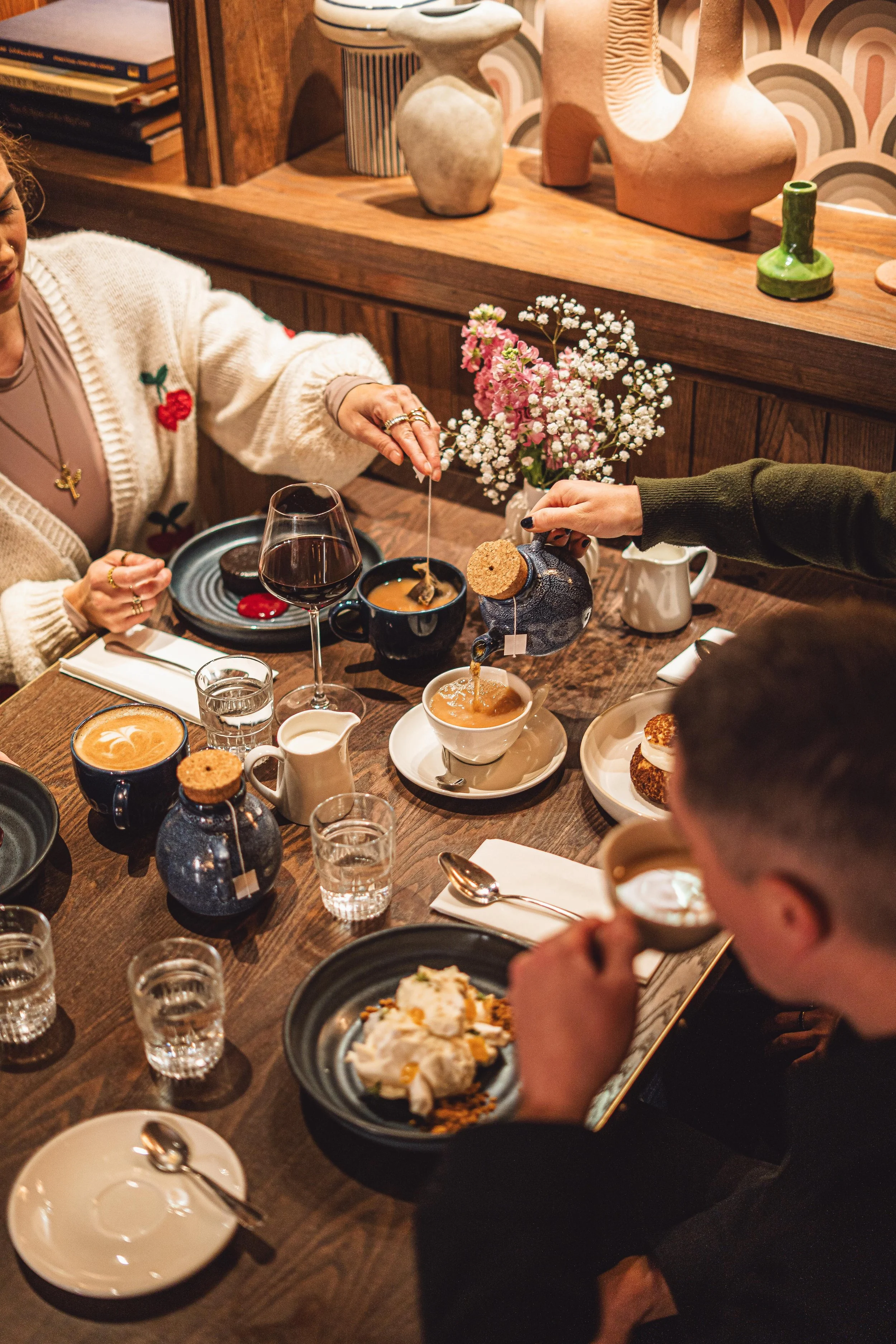 People enjoying coffee and dessert at a cozy wooden table in a cafe, with various cups, jars, flowers, and decorative items on the table and shelves.