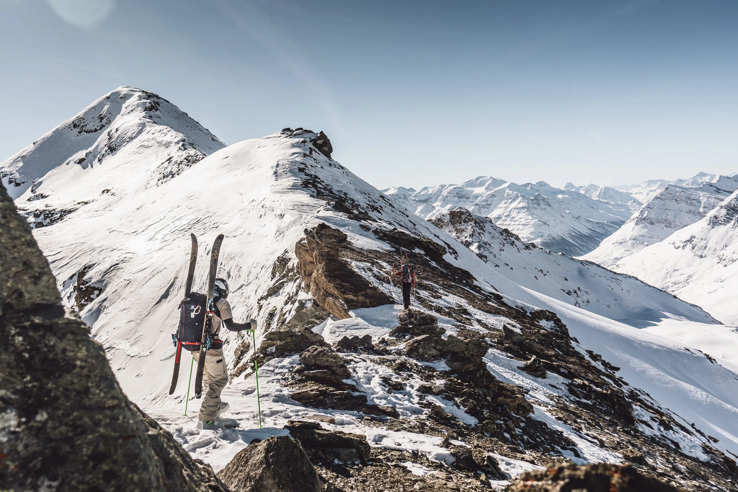 Two skiers with backpacks and ski poles hiking on a snowy mountain ridge with snow-covered peaks in the background.