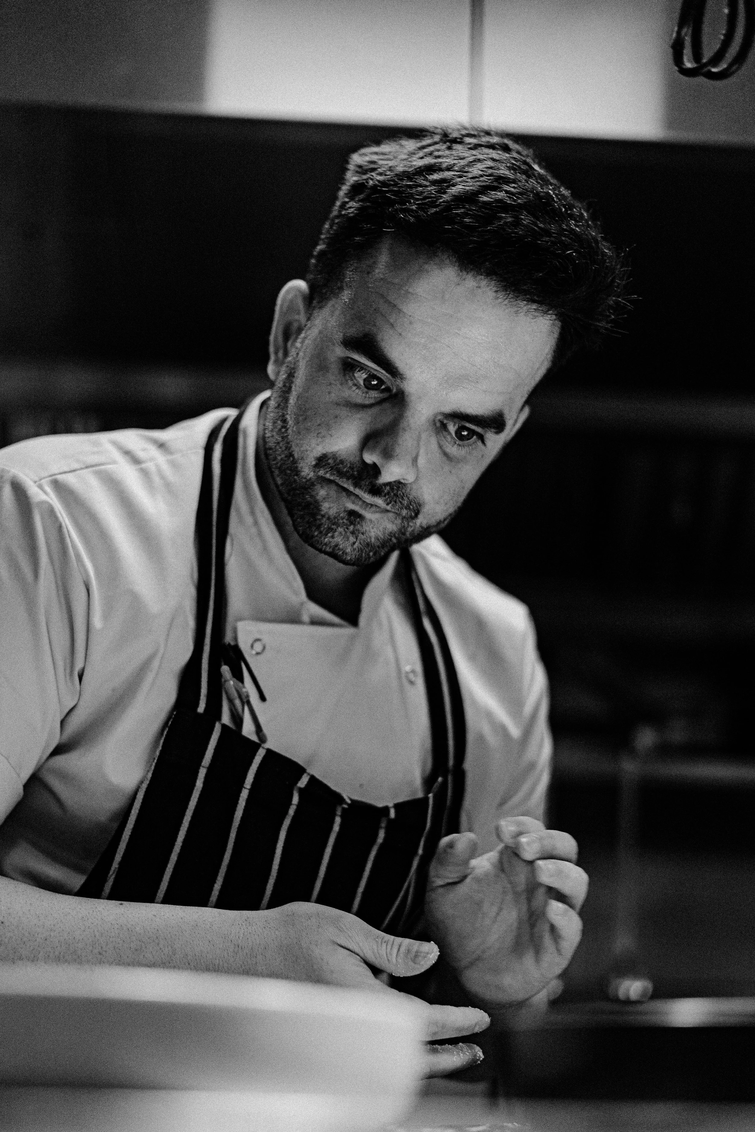 Black and white photo of a male chef in a striped apron young man with facial hair, focused on preparing food in a professional kitchen.
