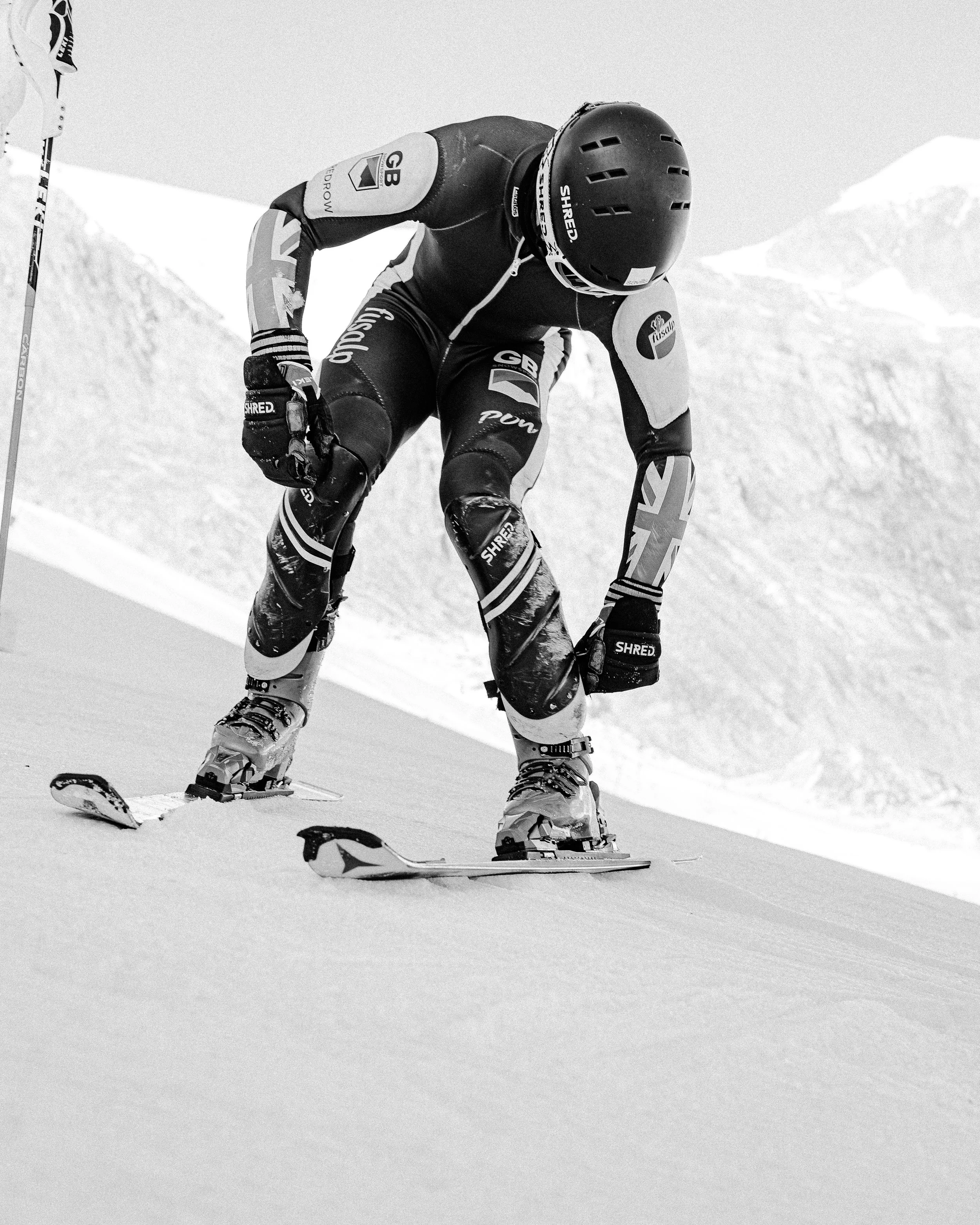 A skier in competitive gear bending down on snow, surrounded by mountainous landscape.