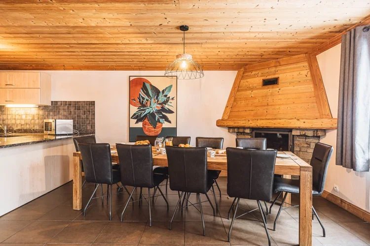 A dining room with a wooden table and eight black chairs in front of a fireplace with a wooden mantel and a large colorful abstract art piece on the wall. There is a hanging light fixture above the table and a window with gray curtains.