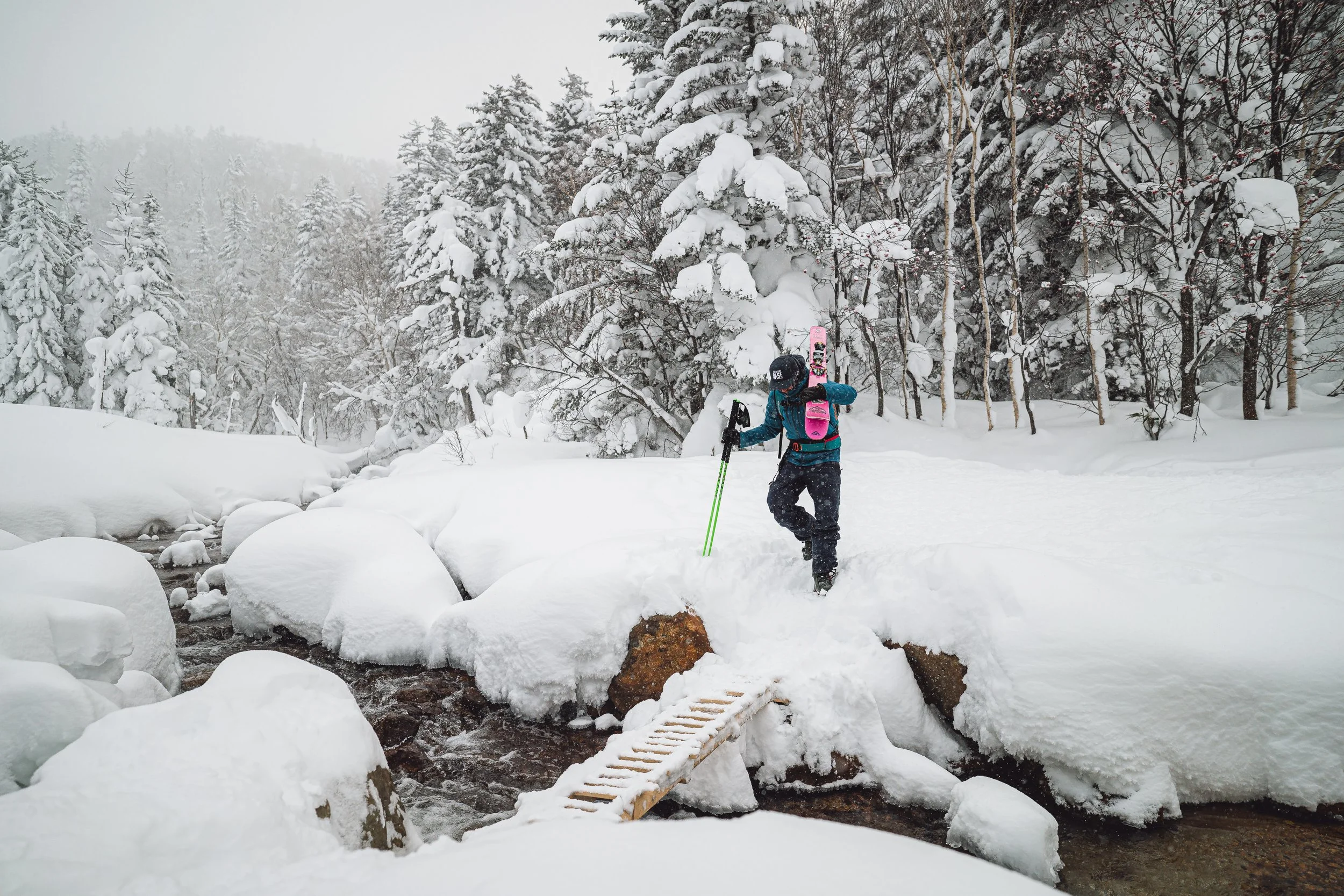 A person dressed in winter gear with a backpack and a helmet crossing a snow-covered creek using a small wooden bridge in a forest during winter.