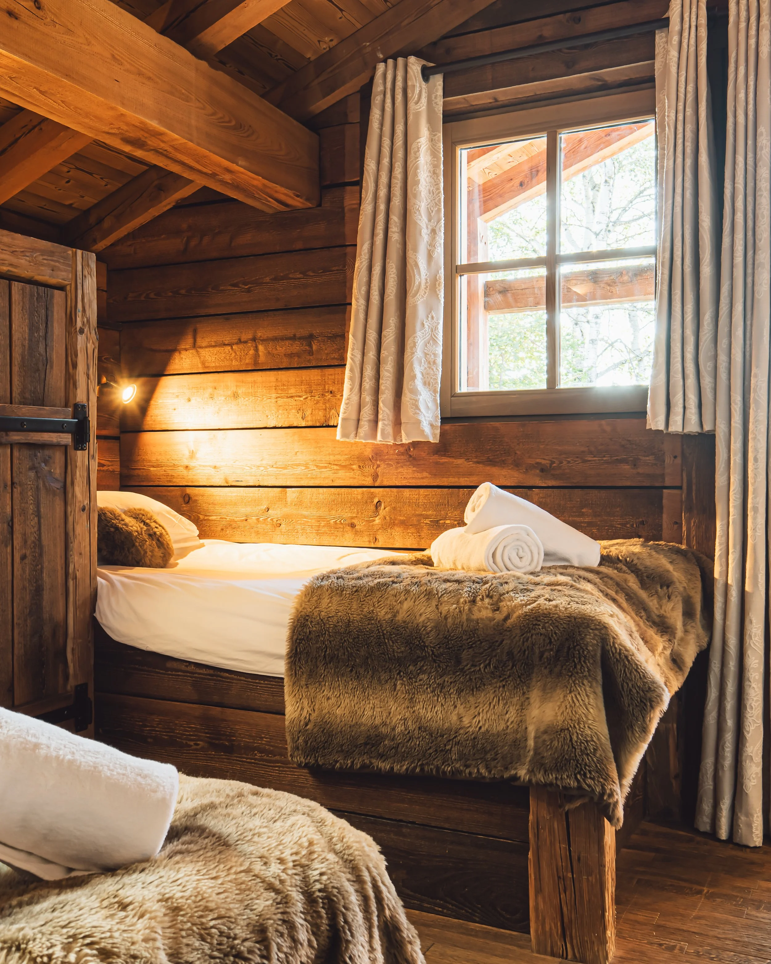 Cozy rustic bedroom with a wooden bed framed by a fur blanket, rolled towels on the bed, and a window with cream patterned curtains letting in natural light.
