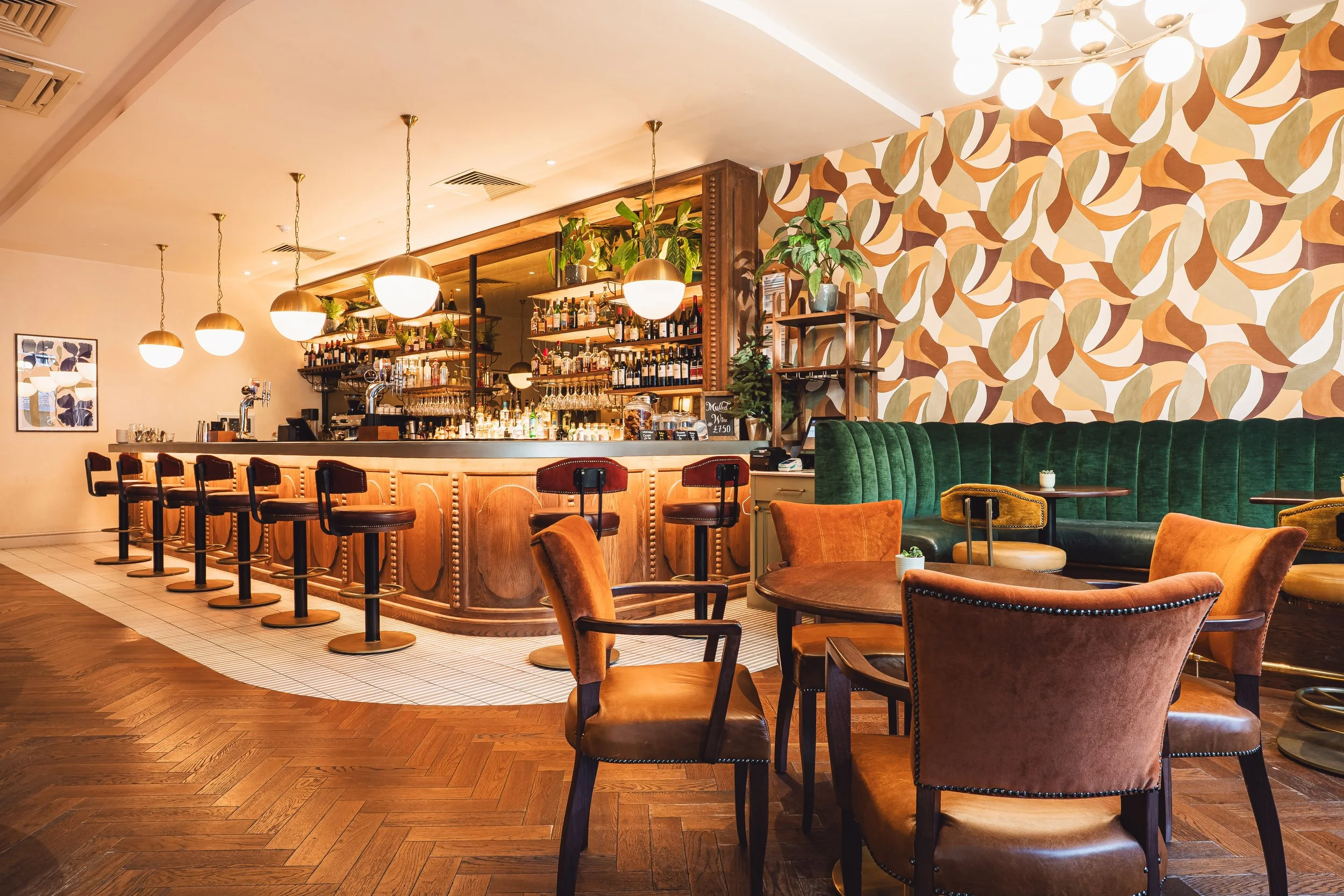 Interior of a stylish bar with wooden decor, bar stools, and cozy seating area with green and brown velvet chairs and patterned wallpaper.
