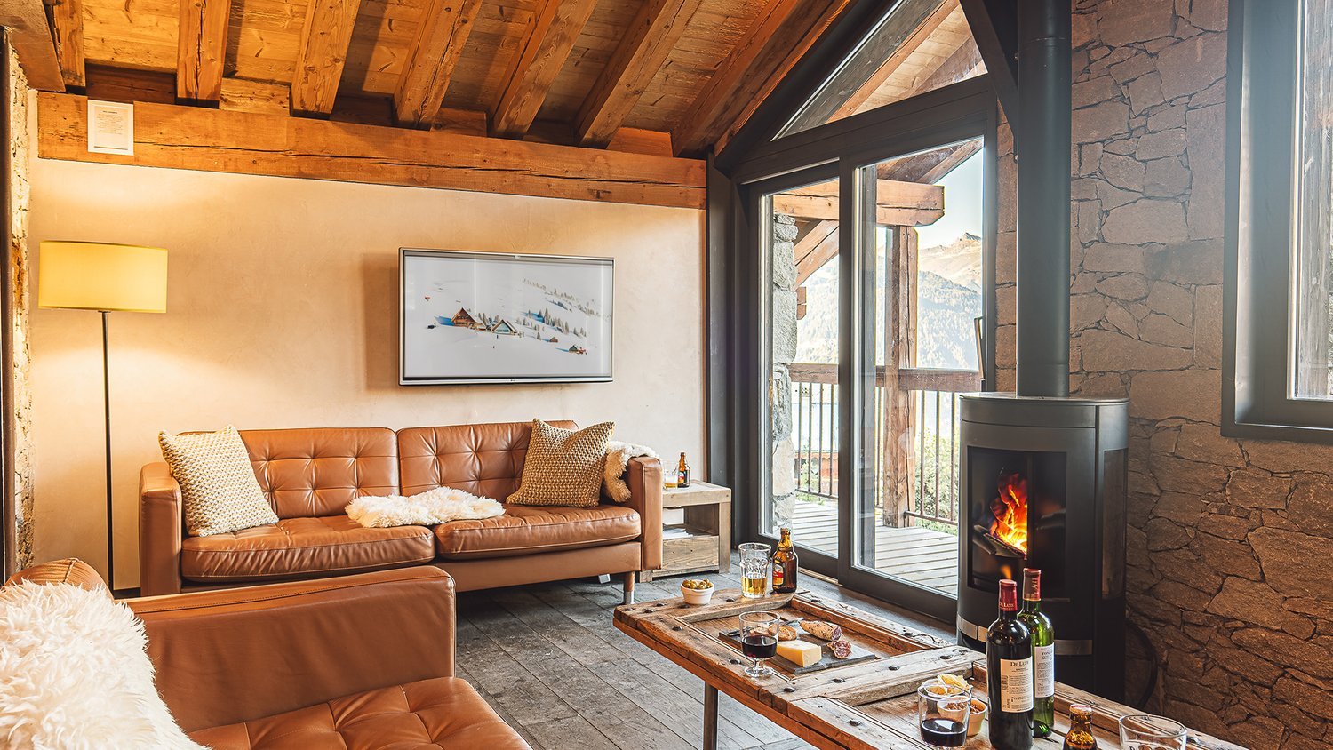 Cozy living room with a brown leather couch, a rocking chair, and a coffee table with drinks and snacks. A fireplace is burning in the corner, with large windows showing a mountain landscape outside.