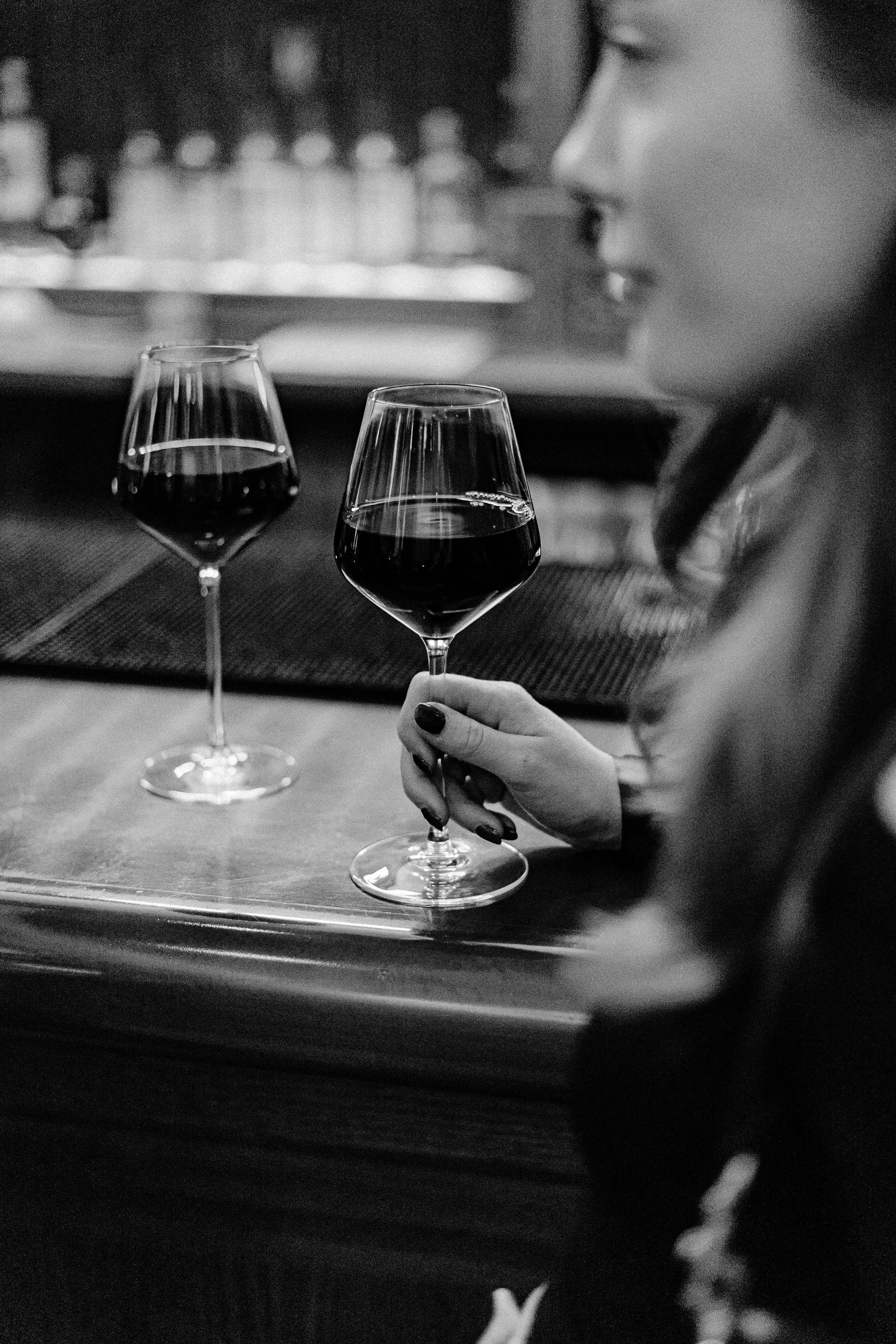 A woman with dark painted nails holding a glass of red wine at a bar, with another glass of red wine and a woman’s face visible in the background.