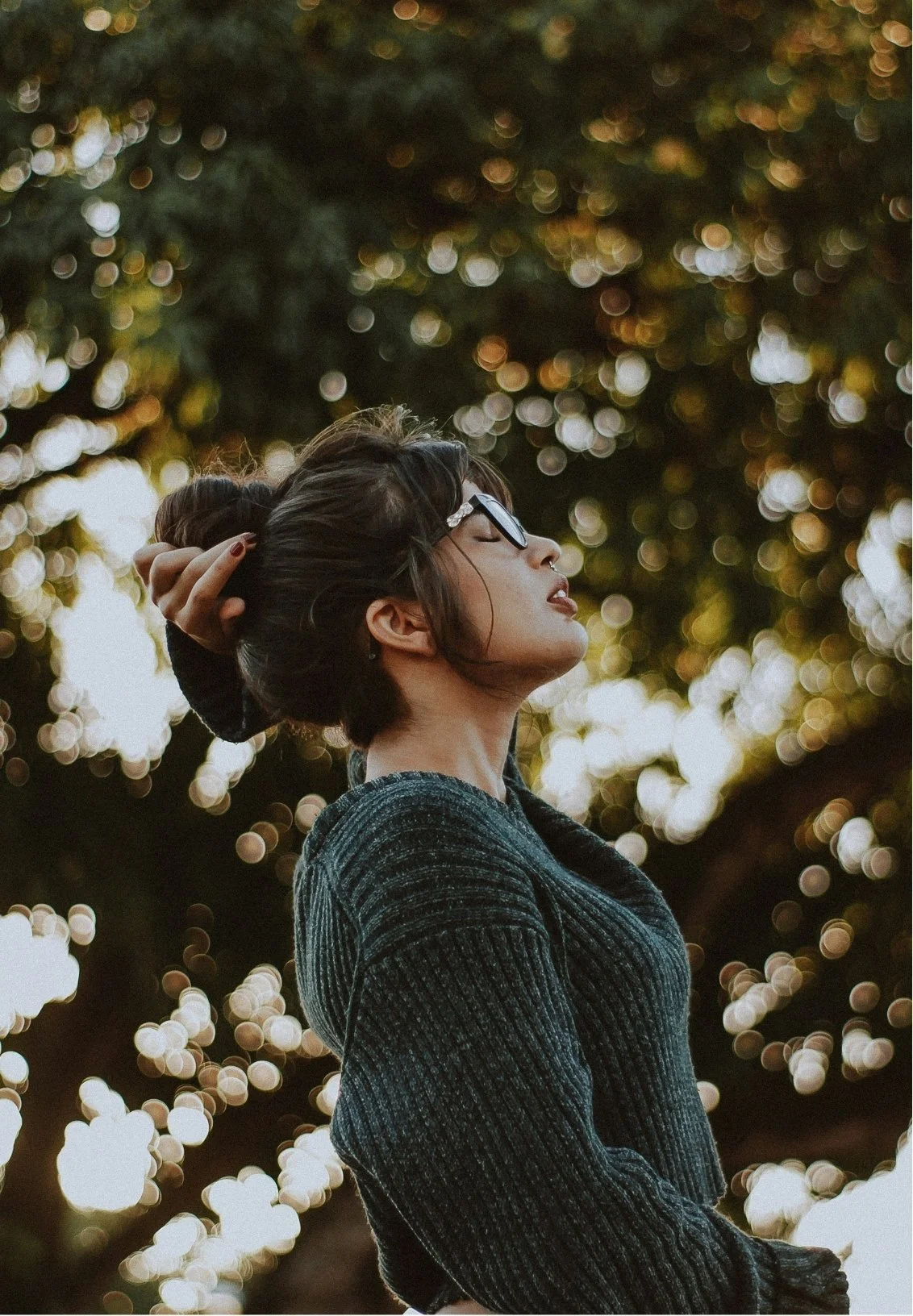 A woman with short dark hair, wearing glasses and a dark sweater, stands outdoors with her eyes closed and head tilted back, touching her hair, with blurred trees and sunlight in the background.