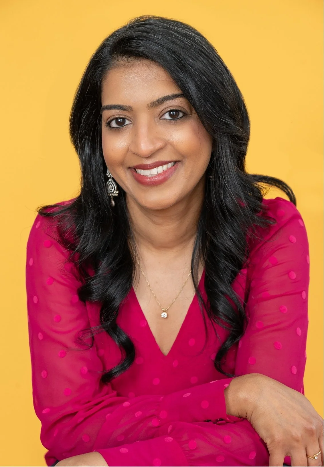 Dr. Shaayestah Merchant, a therapist in Denver, CO wearing a pink polka dot dress, jewelry including earrings and a necklace, smiling, against a yellow background.