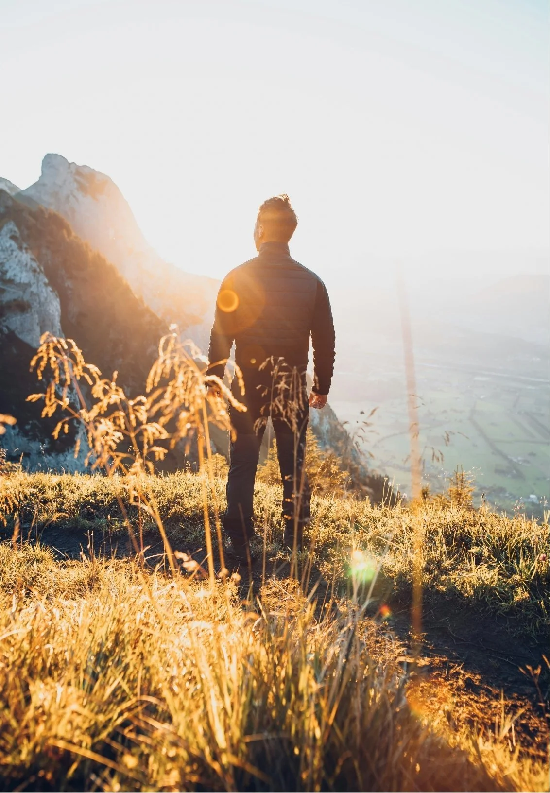 A person standing on a grassy hilltop at sunset, overlooking a mountainous landscape with peaks and a valley.