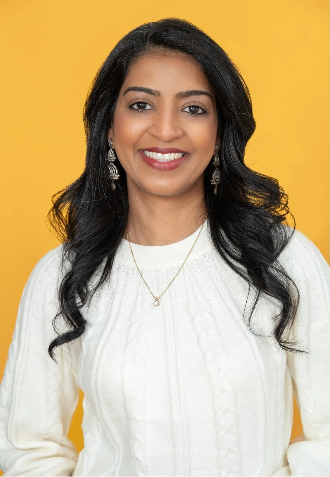 Dr. Shaayestah Merchant, a therapist in Denver, CO, smiling, wearing a white knit sweater, silver earrings, gold necklace, against a yellow background.
