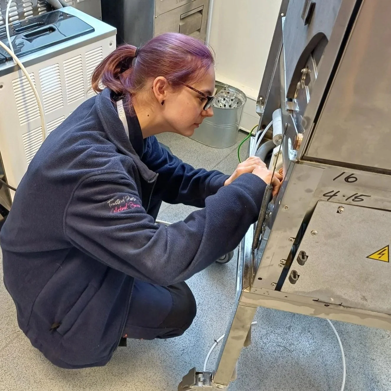 A woman with purple hair in a ponytail, wearing glasses and a dark jacket, is crouching and working on a piece of machinery with various cables and metallic parts, in an industrial or laboratory setting.
