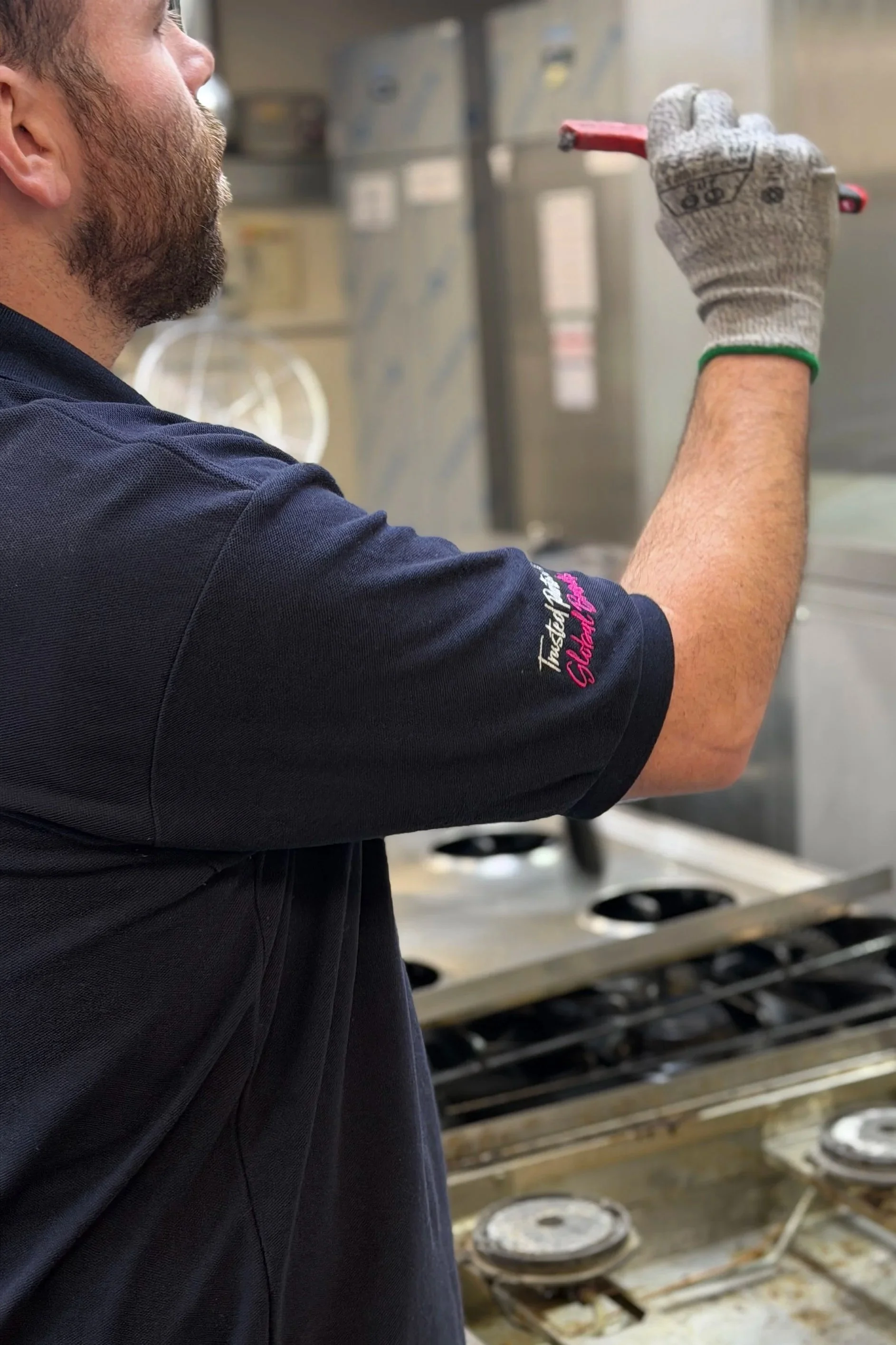 A worker with a beard wearing a dark shirt and gloves, holding a red marker, in a commercial kitchen with a stovetop and cooking equipment in the background.