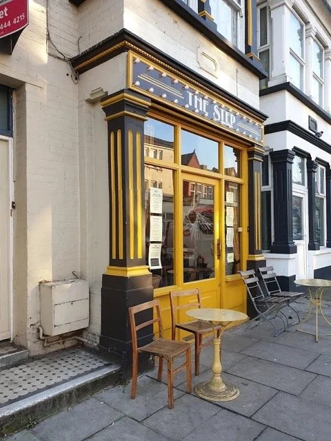 Exterior of a storefront named 'The Step' with black and gold columns, large windows, and outdoor seating including wooden chairs and small round tables on the sidewalk.