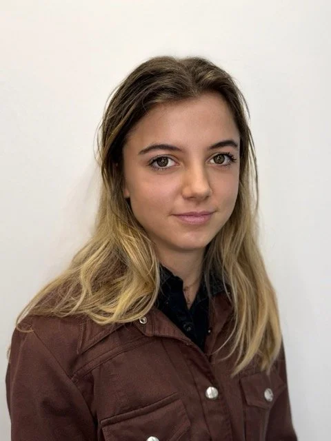 A young woman with long, wavy blonde hair, light skin, and gray eyes, wearing a brown jacket over a black shirt, standing against a plain white wall.
