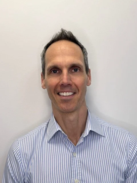 Portrait of a smiling man with short dark hair, wearing a light blue and white striped dress shirt, standing against a plain white wall.