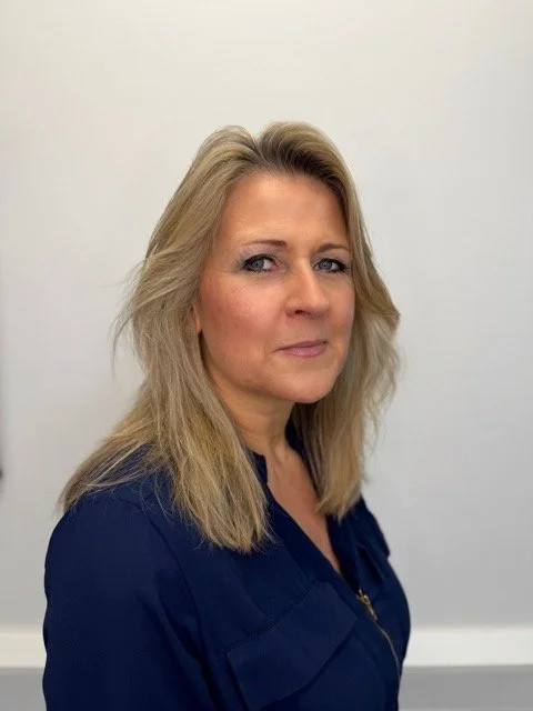 A woman with blonde hair and blue eyes looking at the camera against a plain white wall.