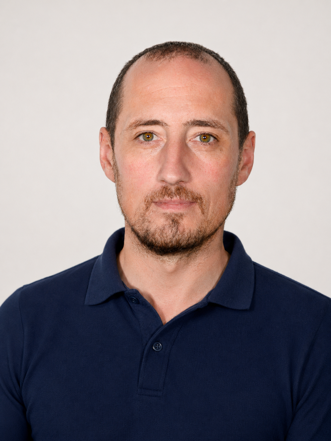 Headshot of a man with short brown hair, light skin, and a beard, wearing a dark blue polo shirt, against a plain background.