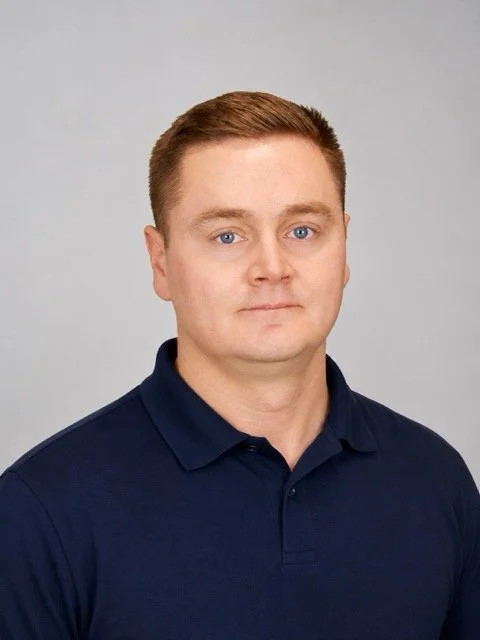 Portrait of a man with short reddish-brown hair, light skin, wearing a navy blue polo shirt, against a plain light gray background.