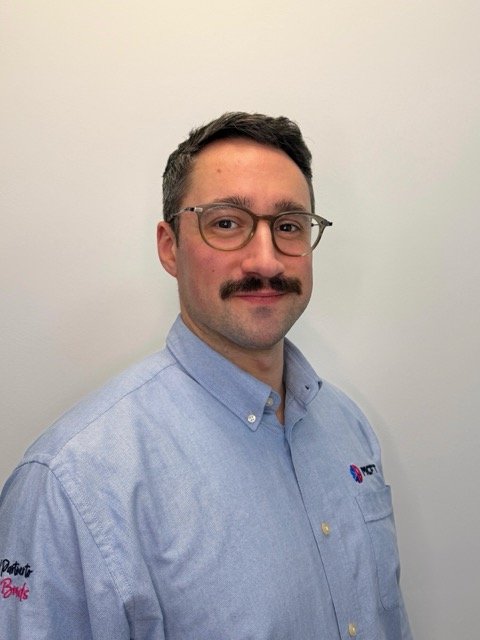 A man with glasses and a mustache wearing a light blue collared shirt with logos, standing against a plain white wall.