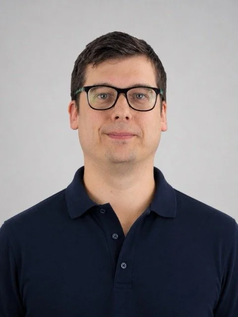 A man with short dark hair and glasses wearing a navy blue polo shirt, standing against a plain light gray background.