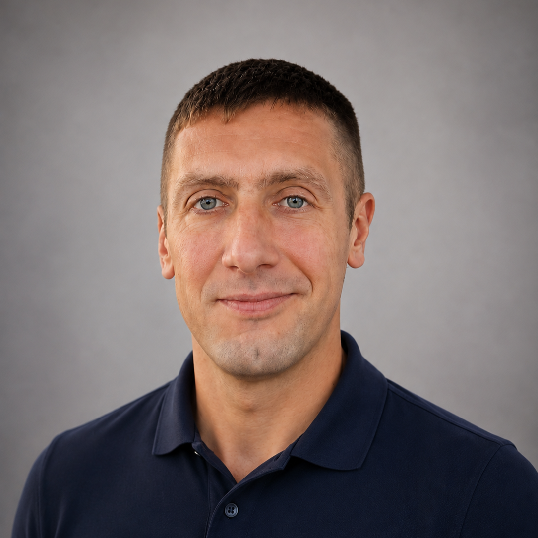 Portrait of a man with short brown hair, blue eyes, wearing a navy blue polo shirt, against a plain gray background.