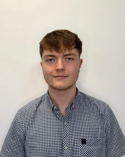 A young man with light skin, short brown hair, and blue eyes, wearing a black and white checkered button-up shirt, standing against a plain white background.