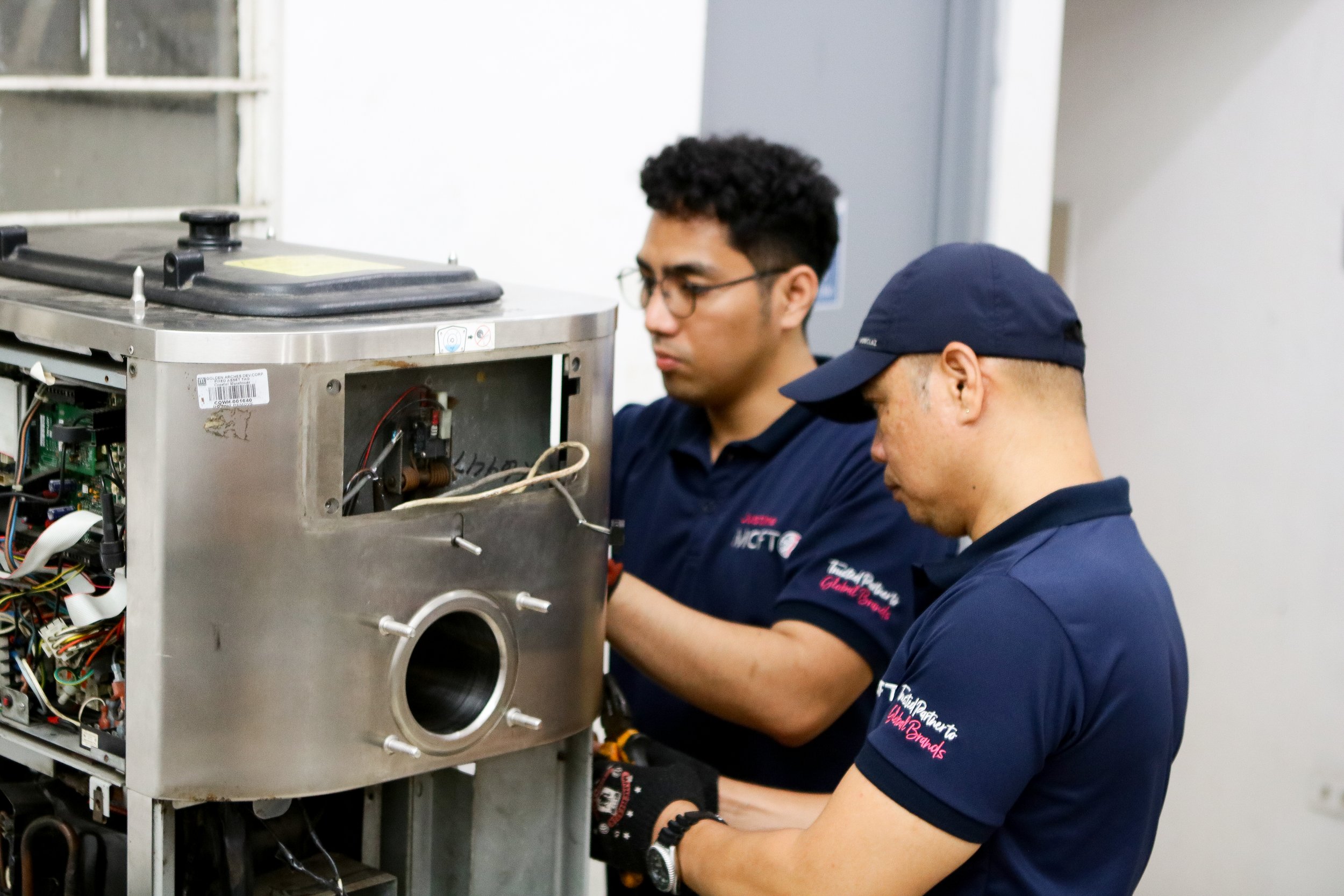 Two technicians working on a piece of electronic equipment, with one of them wearing glasses and the other wearing a cap, both focusing on the device's components.