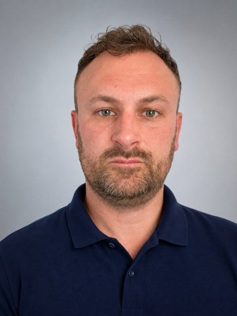 A man with short, curly brown hair, a beard, and blue eyes, wearing a navy blue polo shirt, standing against a light gray background.
