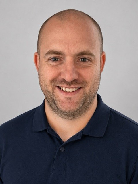 Smiling man with a shaved head and beard wearing a navy blue polo shirt against a plain light gray background.