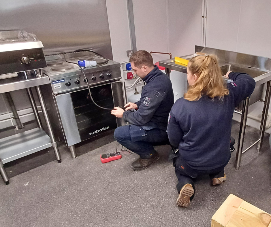 Two firefighters working on a stove, one holding a tool and the other operating a sink in a commercial kitchen.