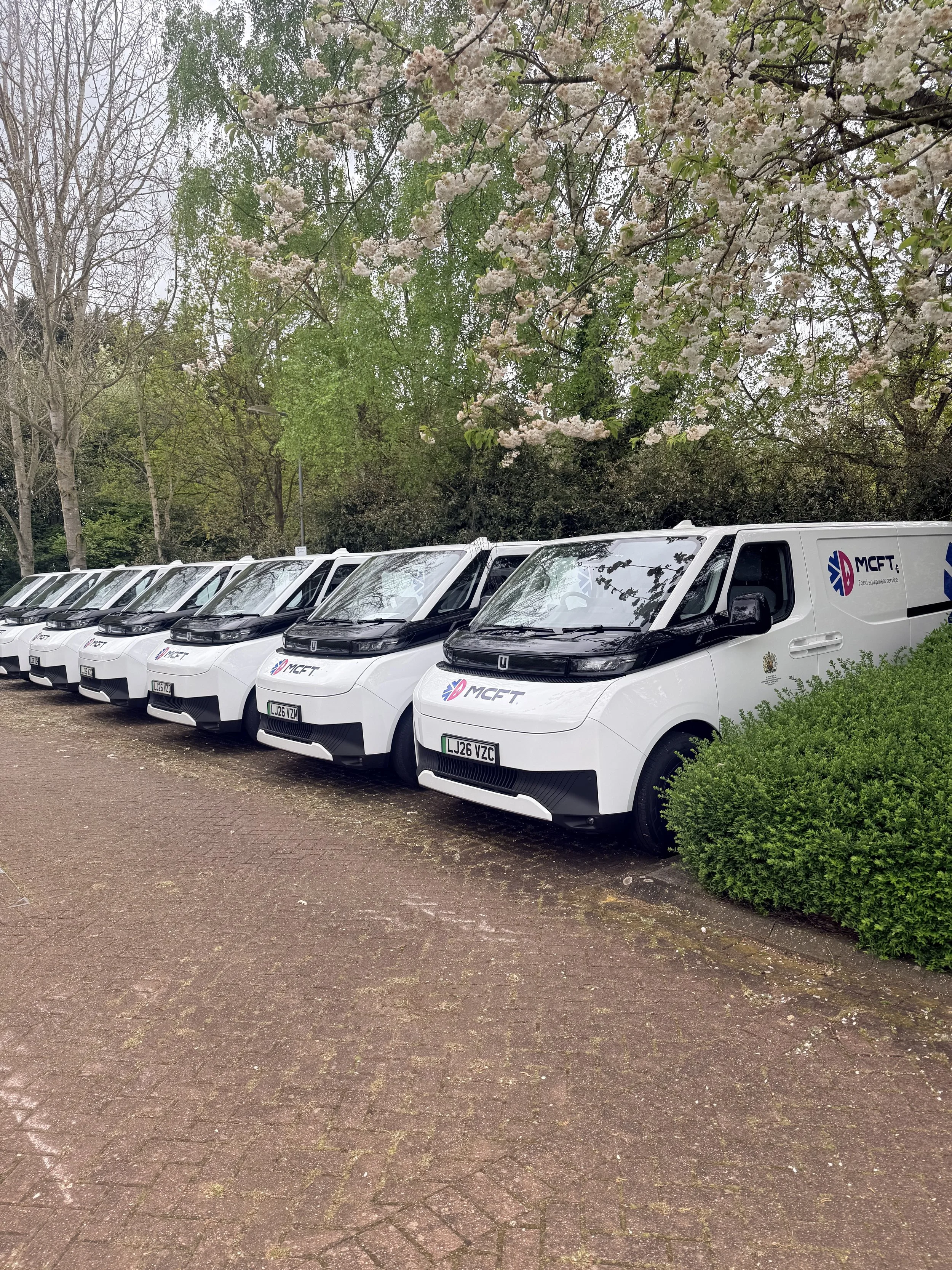 Line of white delivery vans with the logo 'MCFT' parked on a brick-paved area, with trees bearing blossoms and green foliage in the background.